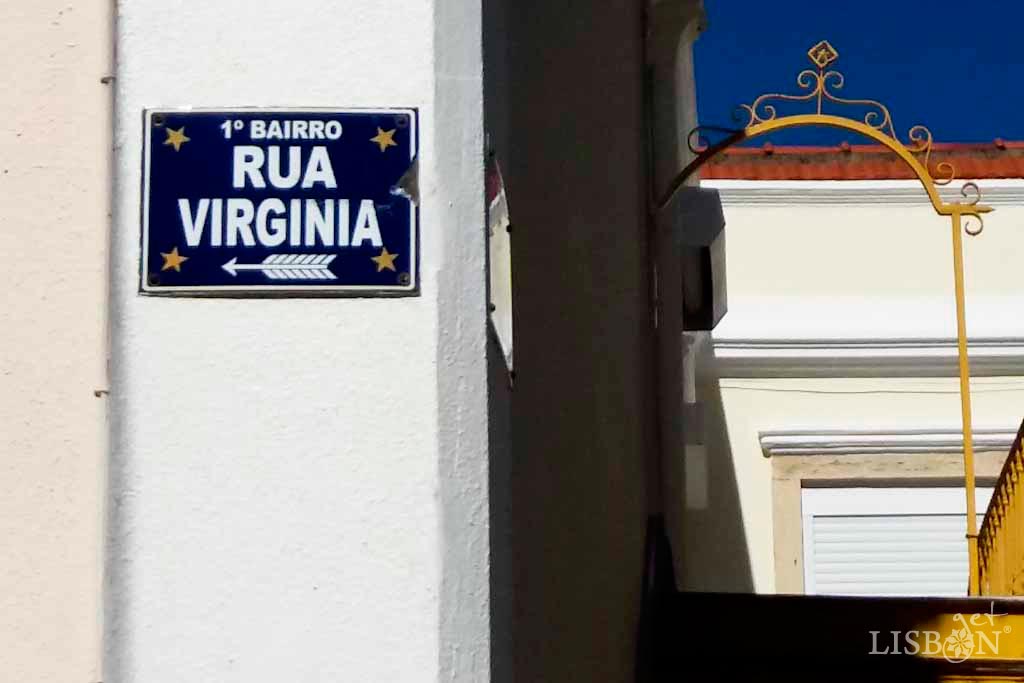 Toponymic plaque of Rua Virgínia. It’s a rectangular metal plaque, painted blue with white letters and each corner has a yellow five-pointed star.