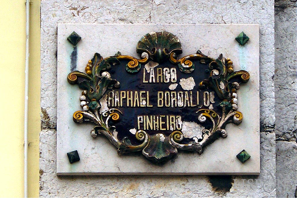 Placa toponímica do Largo Rafael Bordalo Pinheiro, composta por placas cerâmicas recortadas por volutas, acantos e concheados coloridos que emolduram o nome Largo Raphael Bordallo Pinheiro.